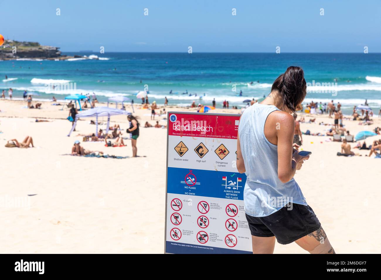 Bondi Beach Sydney 2023, junge brünette Frau, die sms auf ihrem Handy schrieb, Bein- und Armtätowierungen, stand neben dem Bondi Beach Schild, Sydney, NSW, Australien Stockfoto