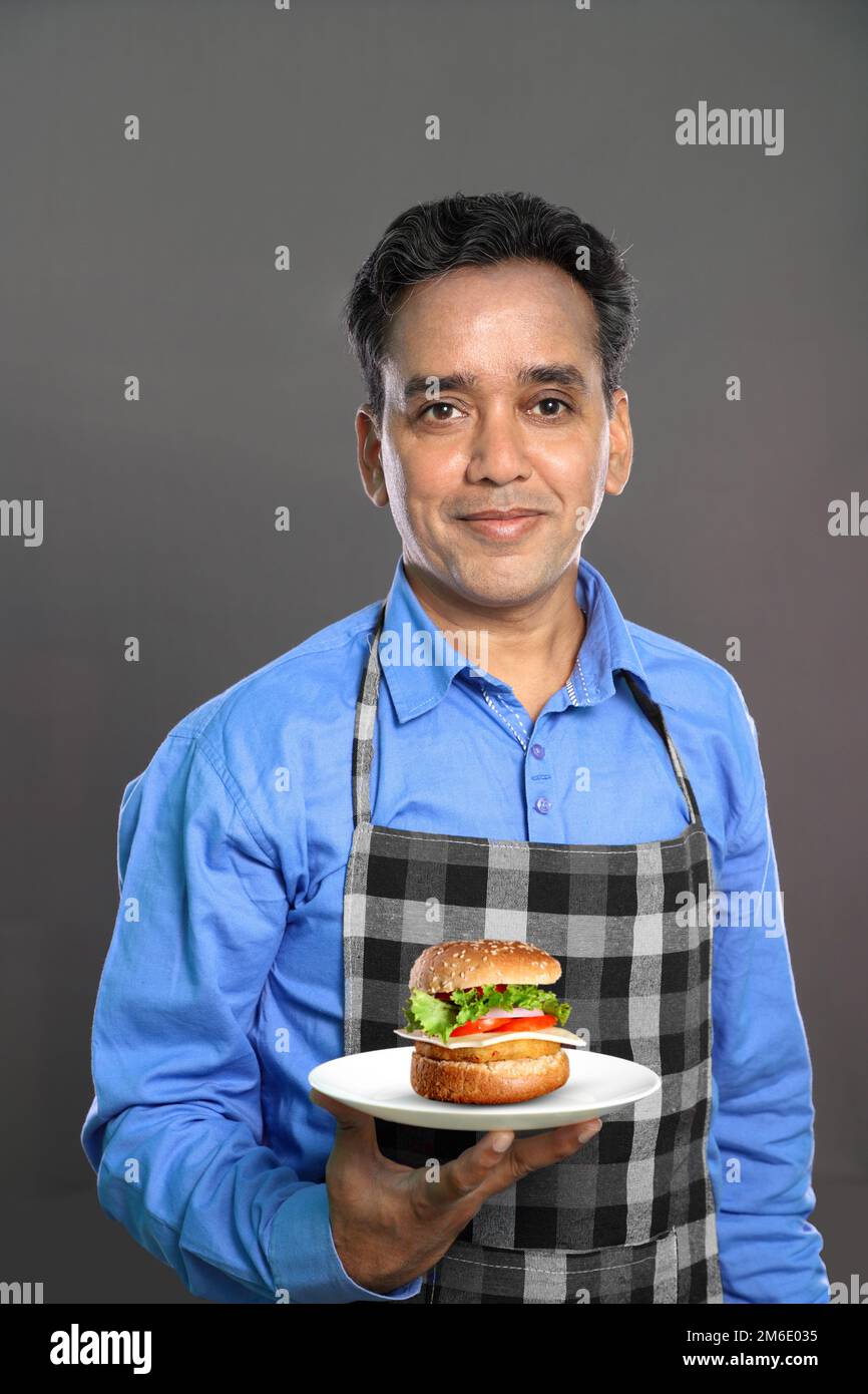 Indischer männlicher Koch mit einem Burger in der Hand auf grauem Hintergrund Stockfoto