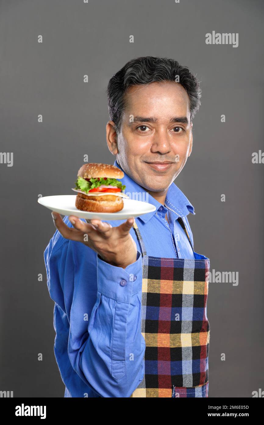 Indischer männlicher Koch mit einem Burger in der Hand auf grauem Hintergrund Stockfoto