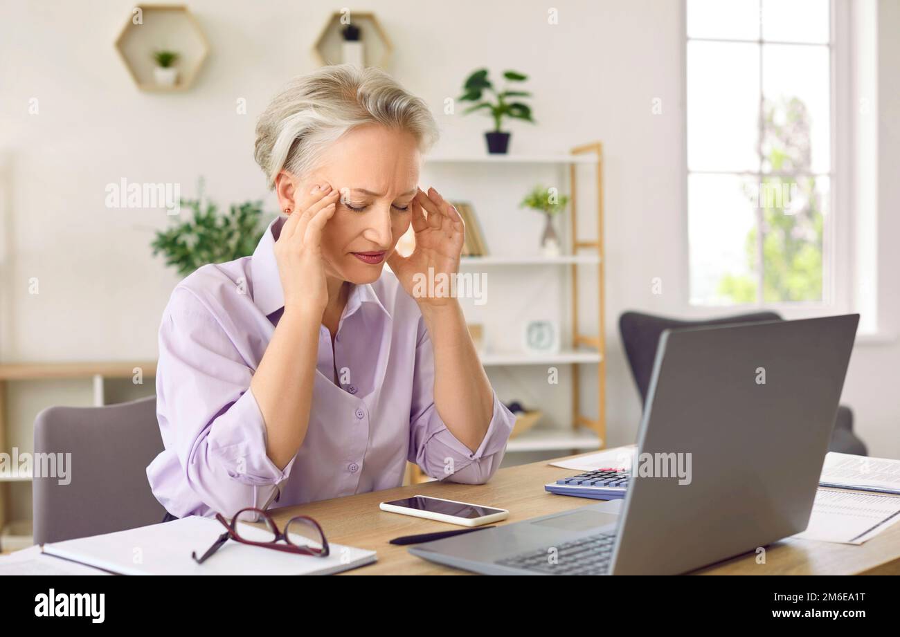 Unglückliche grauhaarige Frau leidet unter Kopfschmerzen und Migräne sitzt am Tisch mit einem Laptop im Büro Stockfoto