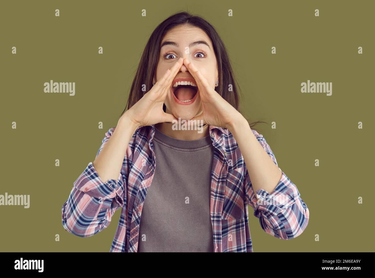 Eine aufgeregte junge brünette Frau in einem karierten Hemd mit den Händen am Mund auf Khaki-Hintergrund. Stockfoto