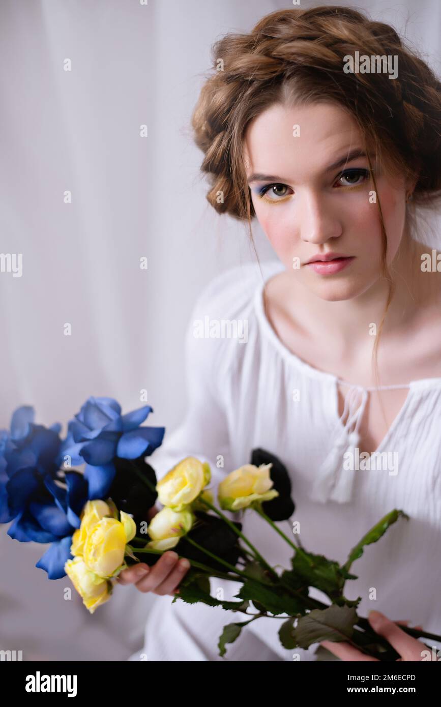 Kaukasische ukrainische Frau, die während des Krieges in einem Kleid und gelb-blauen Blumen auf weißem Hintergrund im Studio sitzt. Konzept - Stoppt den Krieg in der Ukraine. Stockfoto