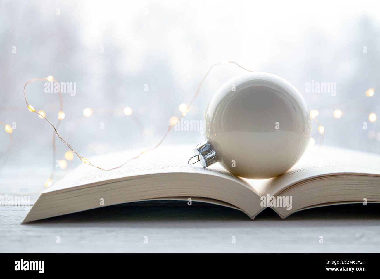 Weiße Weihnachtskugel auf einem offenen Papierbuch mit märchenhaften Lichtern auf dem weißen Tisch, verschneite Waldlandschaft im Hintergrund. Stockfoto