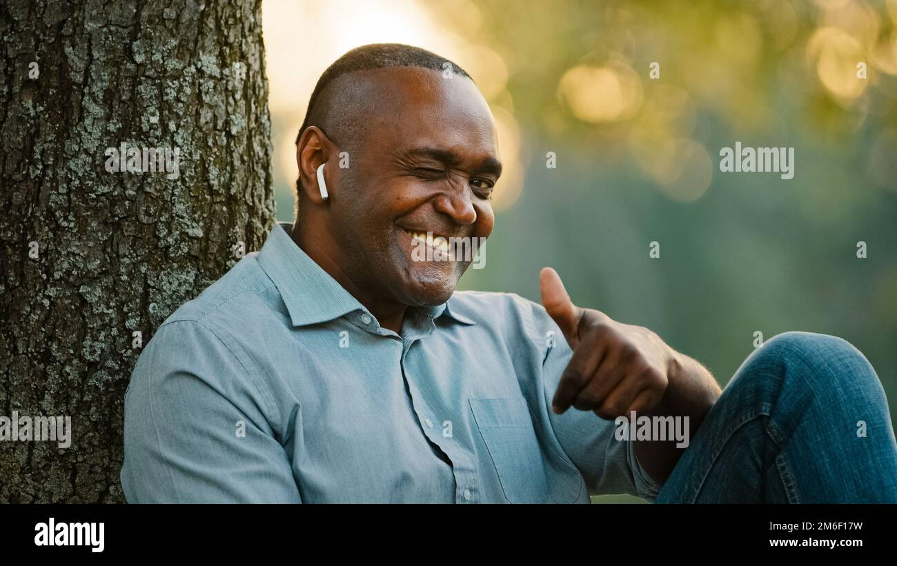 Porträt älterer afroamerikanischer älterer erwachsener Mann mittleren Alters, der im Park unter einem Baum sitzt und sich im Freien ausruht und Musik hört Stockfoto