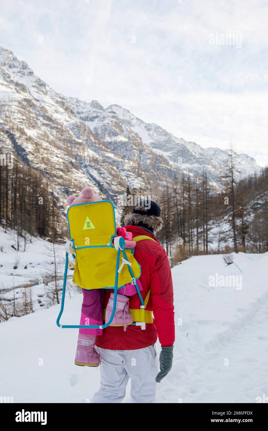 Mann und Frau mit einem Baby in einem Rucksackträger, die auf dem Feld in einer verschneiten Berglandschaft spazieren Stockfoto