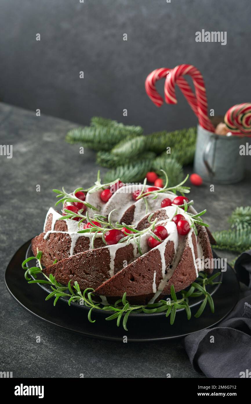 Weihnachtsschokoladenkuchen. Traditioneller Weihnachtskuchen mit weißer Glasur, Preiselbeeren und Rosmarin auf dunklem Hintergrund als Weihnachtsdekoration. Wi Stockfoto