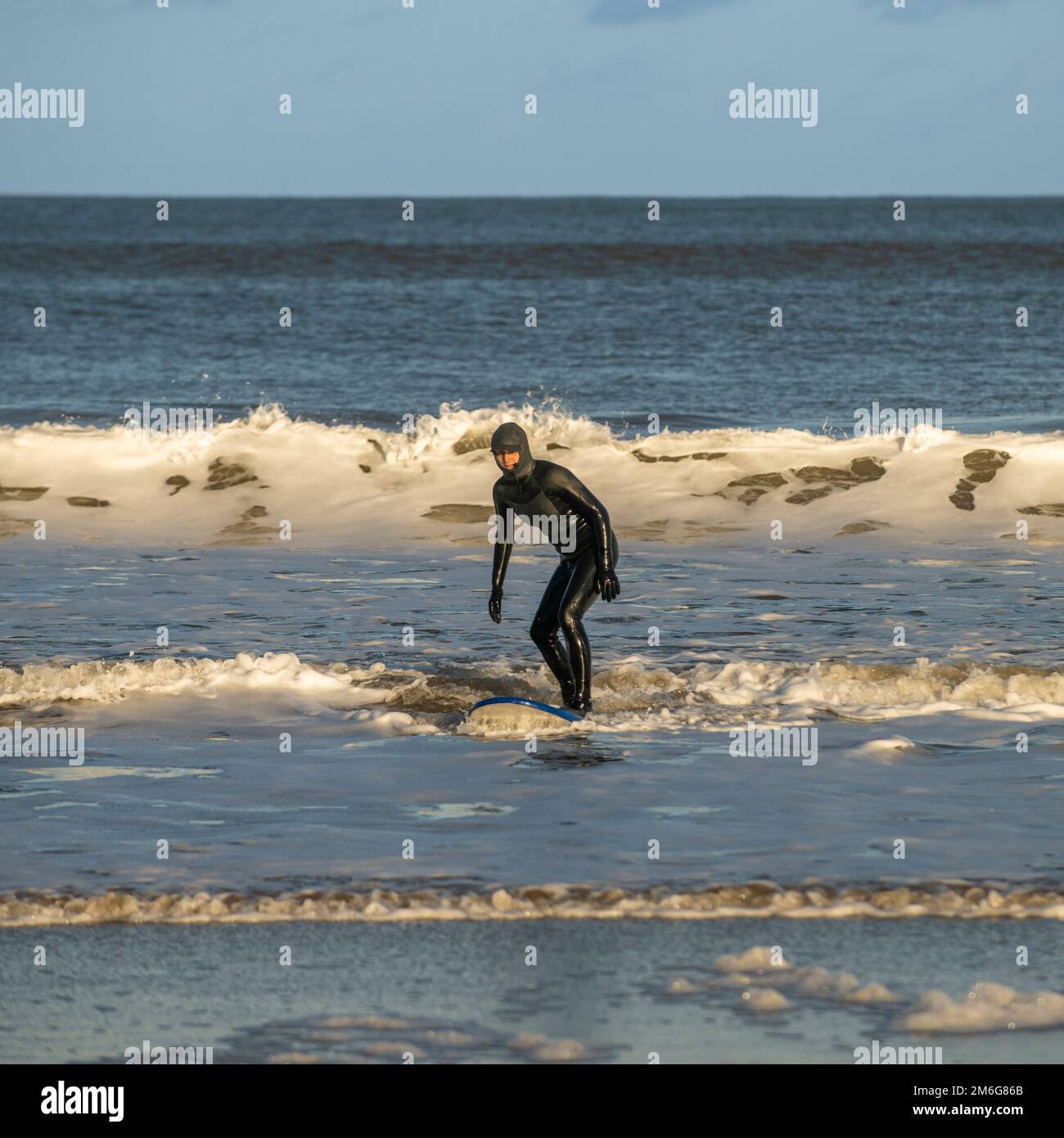Männlicher, kaukasischer Surfer mit schwarzem Neoprenanzug, der auf einem Surfbrett steht, das in Cayton Bay, North Yorkshire, Großbritannien, an Land kommt Stockfoto