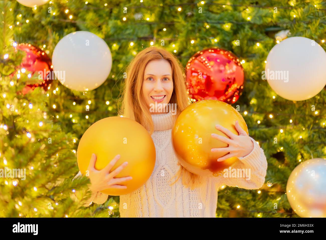 Lächelndes Mädchen mit riesigen Weihnachtsbällen auf dem Hintergrund eines eleganten Weihnachtsbaums. Festliches Poster. Herzlichen Glückwunsch Stockfoto