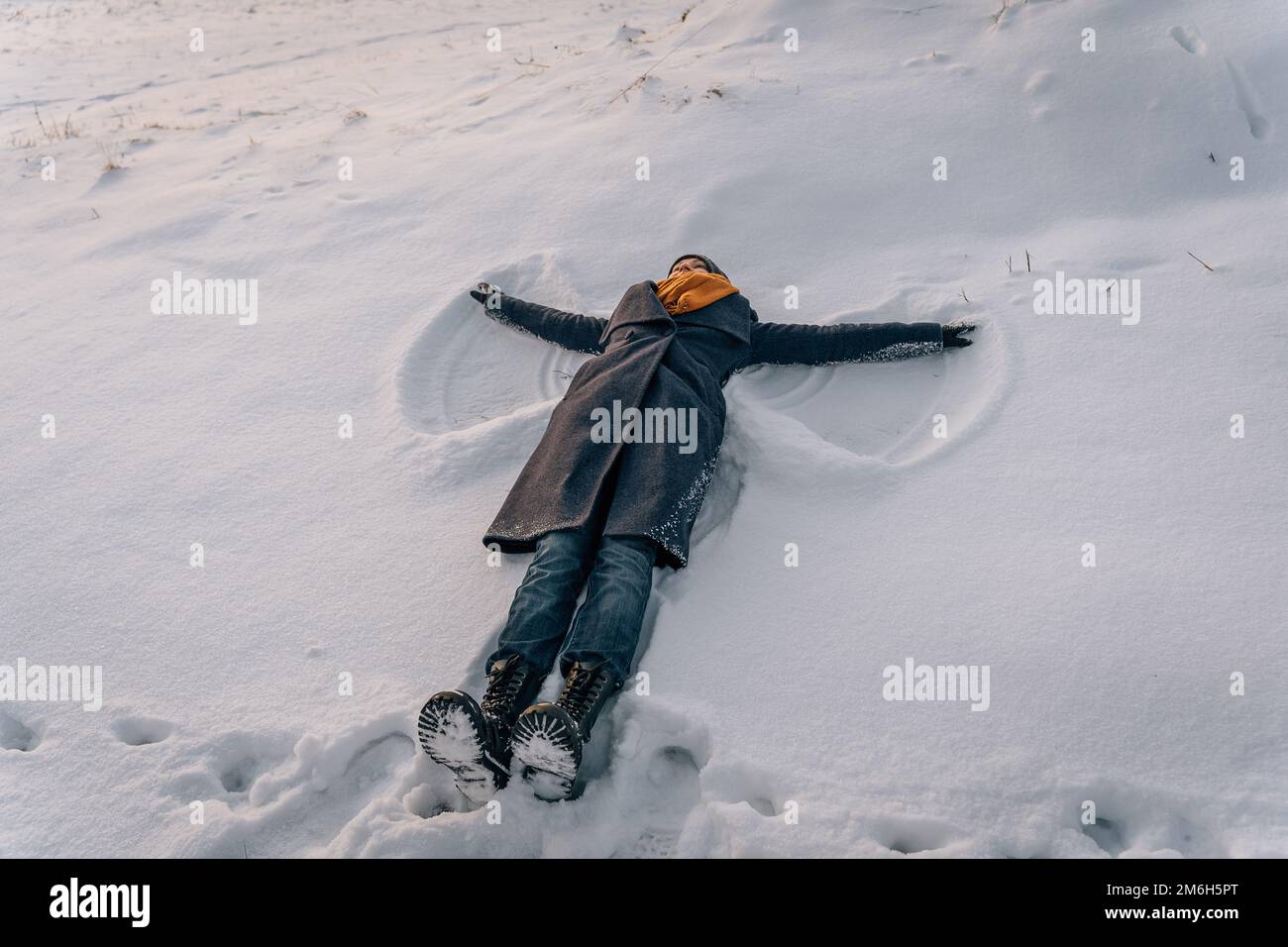 Eine Frau mit Mantel, Schal und Stiefeln liegt auf einem schneebedeckten Feld und macht einen Schneeengel. Stockfoto