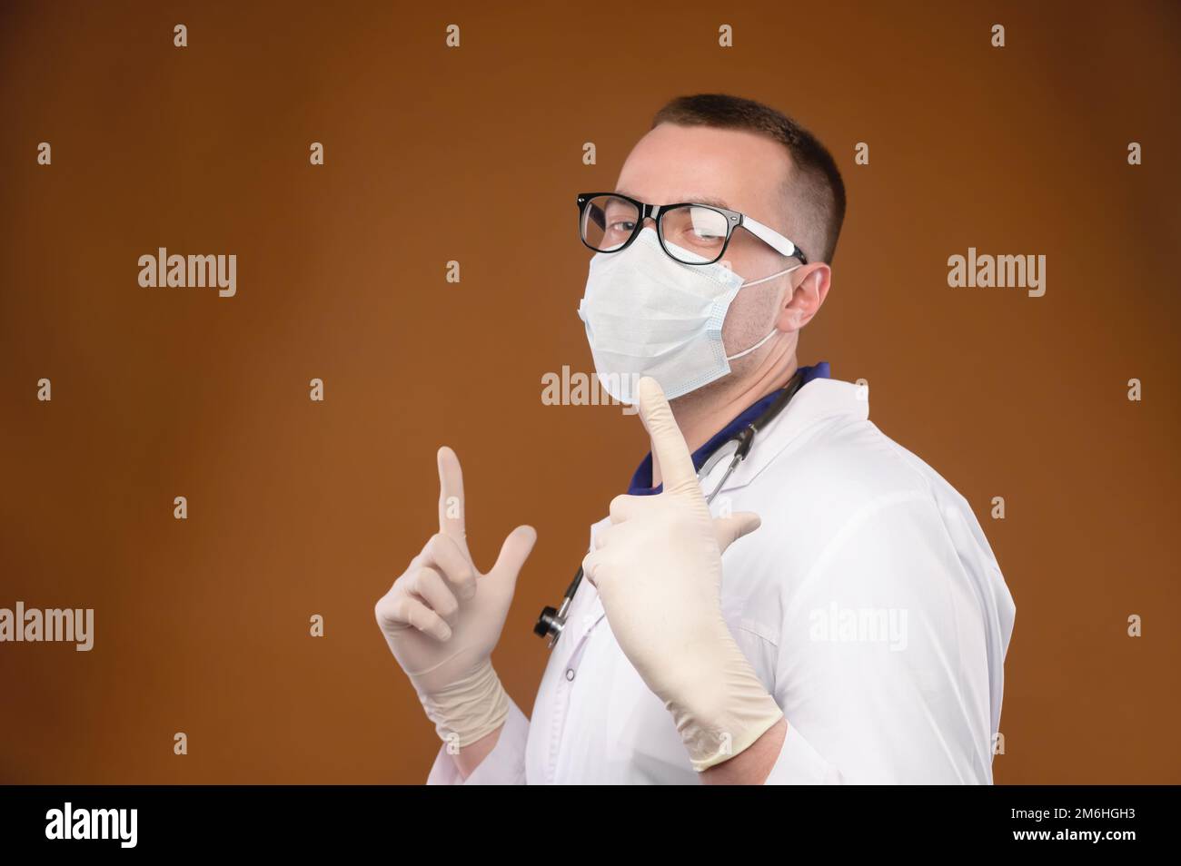 Ein glücklicher weißer Sanitäter in einem weißen Mantel mit Brille und Maske tanzt im Studio und zeigt mit den Zeigefingern nach oben. Stockfoto