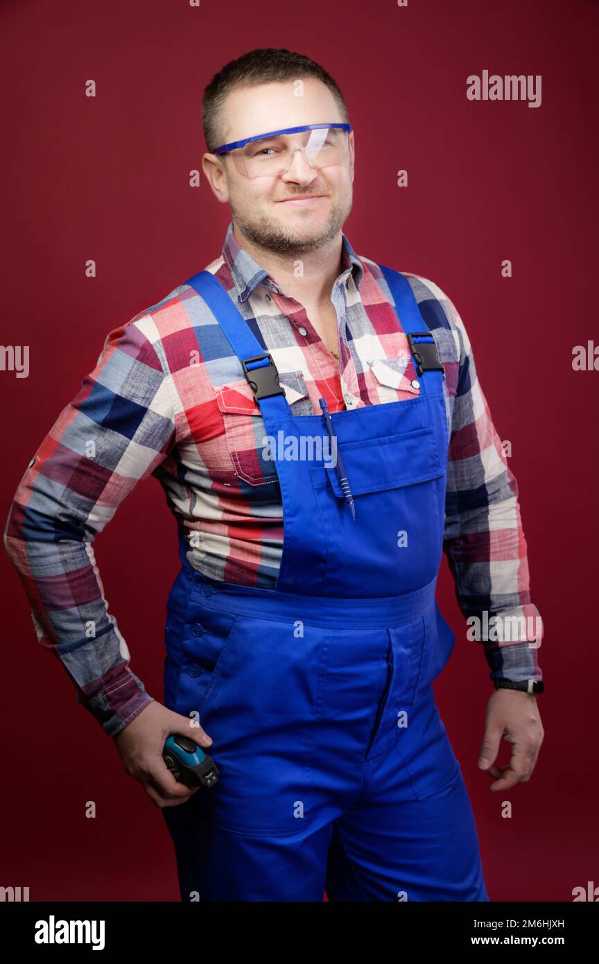 Porträt eines positiv freundlichen kaukasischen männlichen Werkstatters mit Overall und Brille auf dunkelrotem Hintergrund. Lächelnder Blick Stockfoto