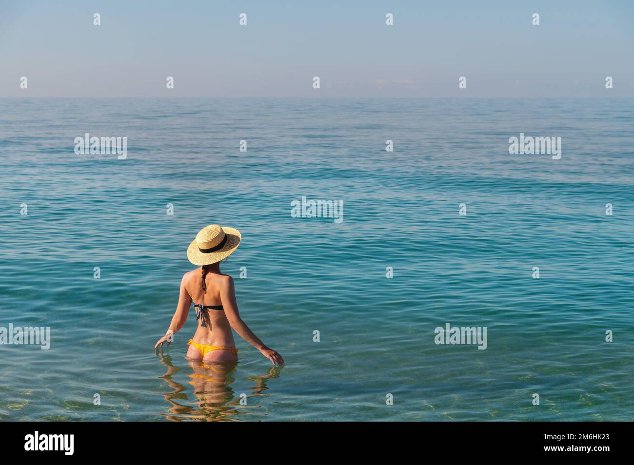 Rückansicht einer schlanken kaukasischen jungen Frau, die in einem Badeanzug und einem Strohhut hüfttief in ruhigem, transparentem Meerwasser steht Stockfoto