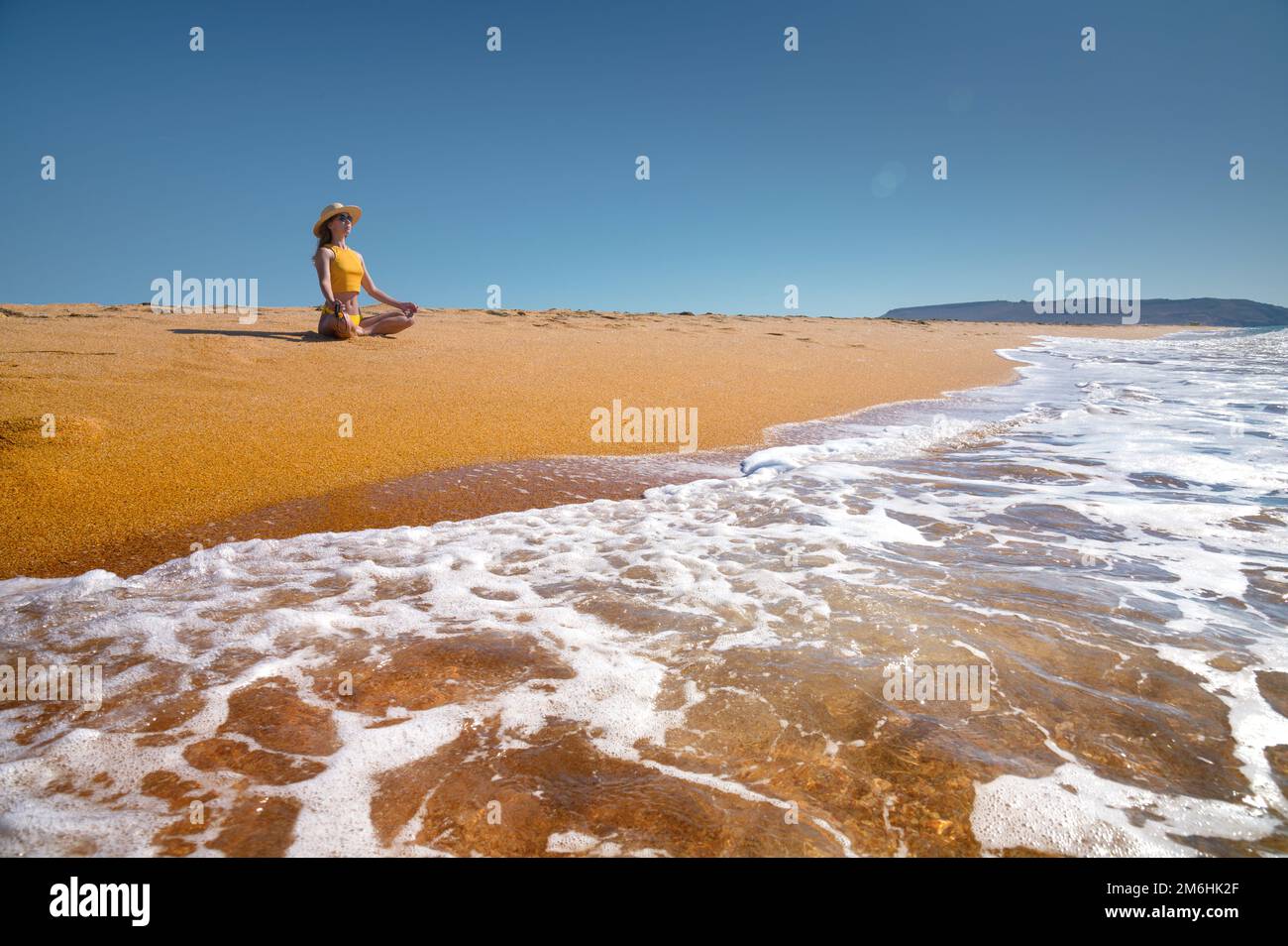 Meditation am Meer an einem sonnigen Tag. Junge weiße Frau in einem gelben Badeanzug sitzt auf dem goldenen Sand in einer Lotusposition Stockfoto