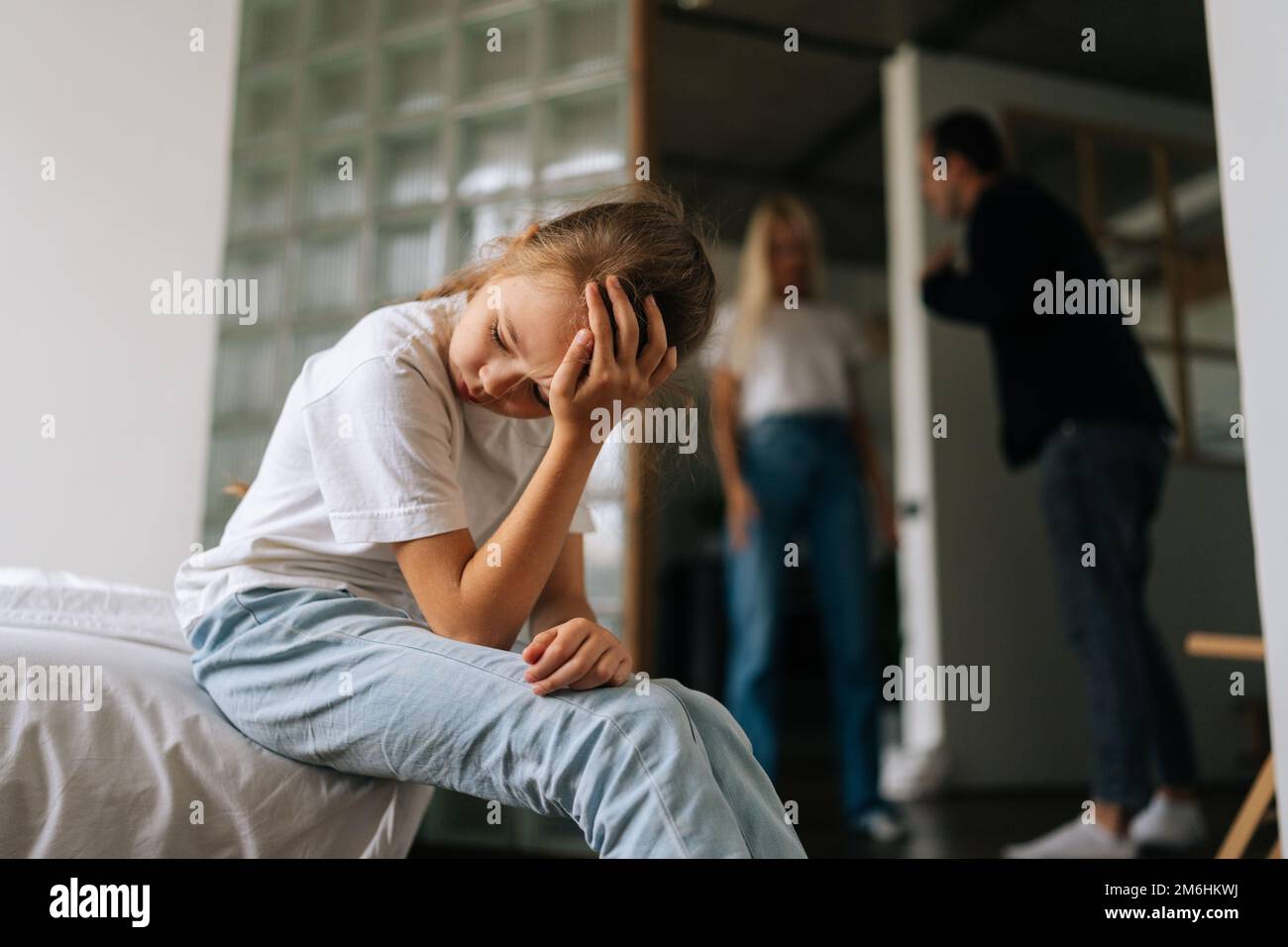 Vorderansicht eines traurigen, einsamen kleinen Mädchens, das wegsieht, weinend leidend auf dem Sofa sitzt, während Eltern sich streiten und im Wohnzimmer streiten Stockfoto
