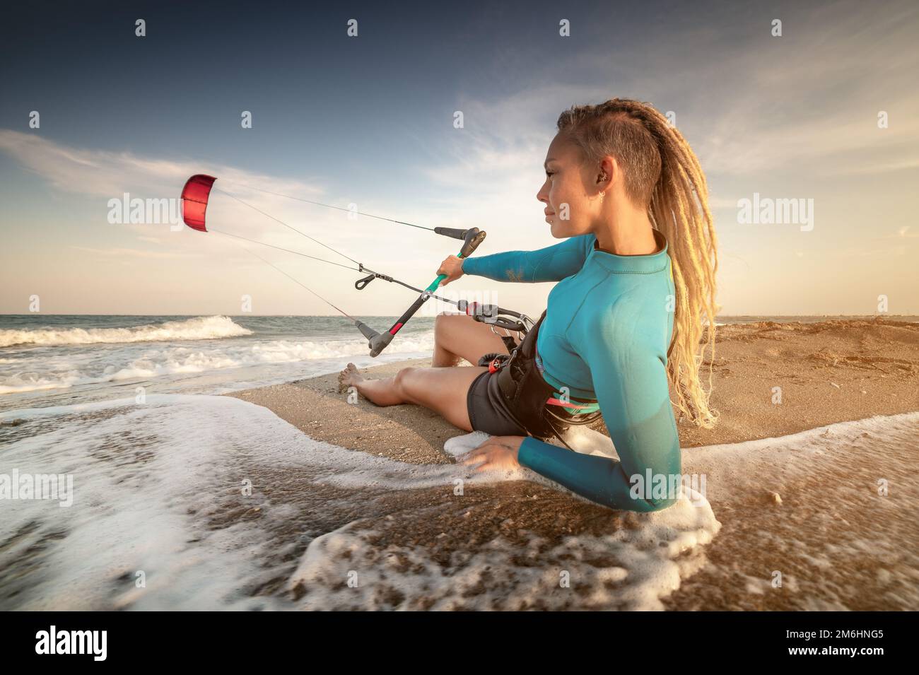 Attraktive weiße Kitesurferin in Neoprenanzug und mit Dreadlocks auf dem Kopf ruht auf einem Sandstrand Stockfoto