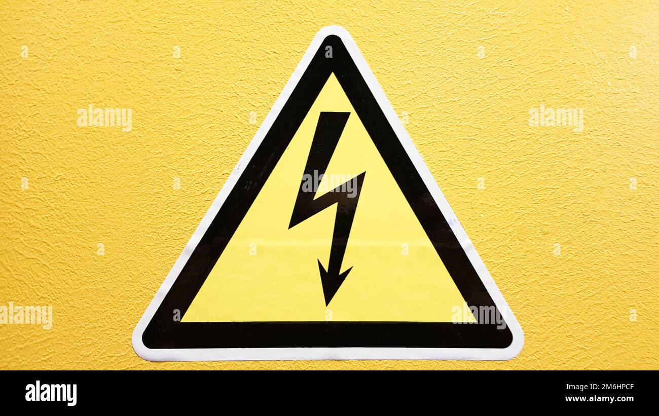Gelbes und schwarzes Sicherheitshinweisschild an einer gelben Wand geklebt. Hochspannungsblitze in einem Dreieck Vorsicht Gefahr Elektrizität dea Stockfoto