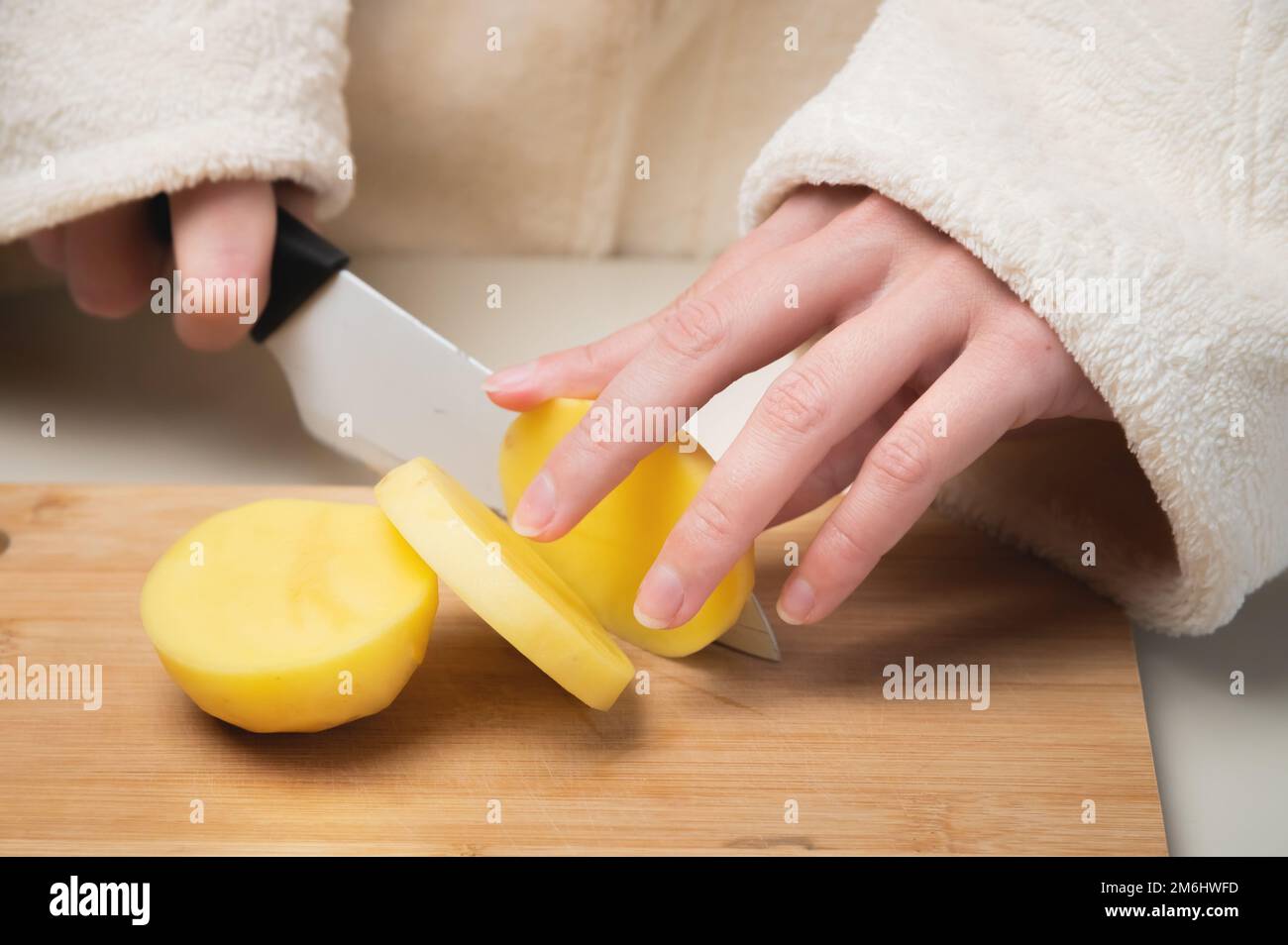 Junge weiße Frau im Bademantel steht und schneidet Kartoffeln auf einem Schneidebrett Stockfoto