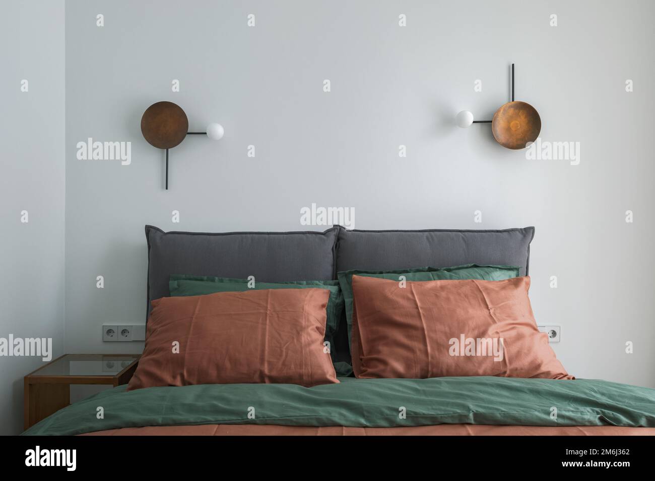 Zimmereinrichtung mit rostfarbener Bettwäsche und Kissen auf einem Bett, grauer Decke, Rattan Nachttisch und Wandleuchte aus Metall. Stockfoto