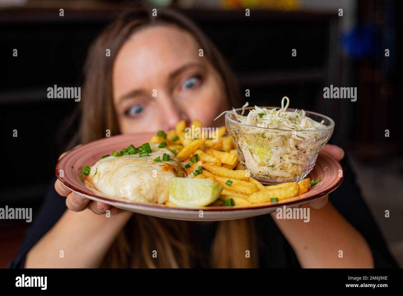 Eine junge Frau hält den Teller mit Pommes Frites, Hühnerfilet und Gemüsesalat in den Händen und starrt mit hungrigen Augen. Servieren von Hauptgericht und Ziernaht Stockfoto