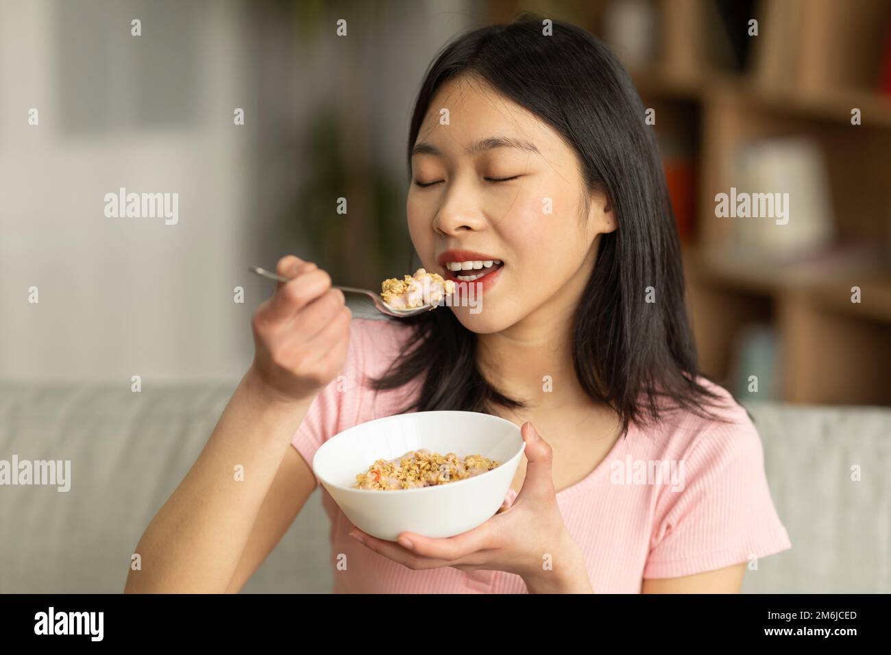 Porträt einer jungen asiatischen Dame, die Müsli mit Milch isst, während sie zu Hause auf dem Sofa sitzt und eine gesunde Mahlzeit genießt Stockfoto