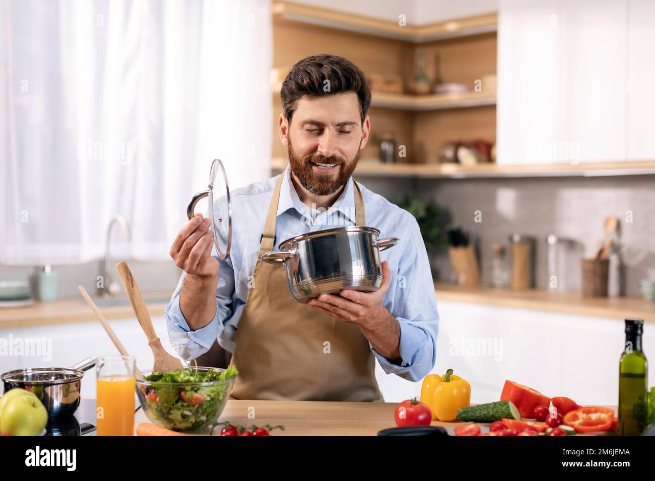 Ein glücklicher weißer, bärtiger Chefkoch in der Schürze, der Essen zubereitet und am Tisch in die Pfanne schaut Stockfoto