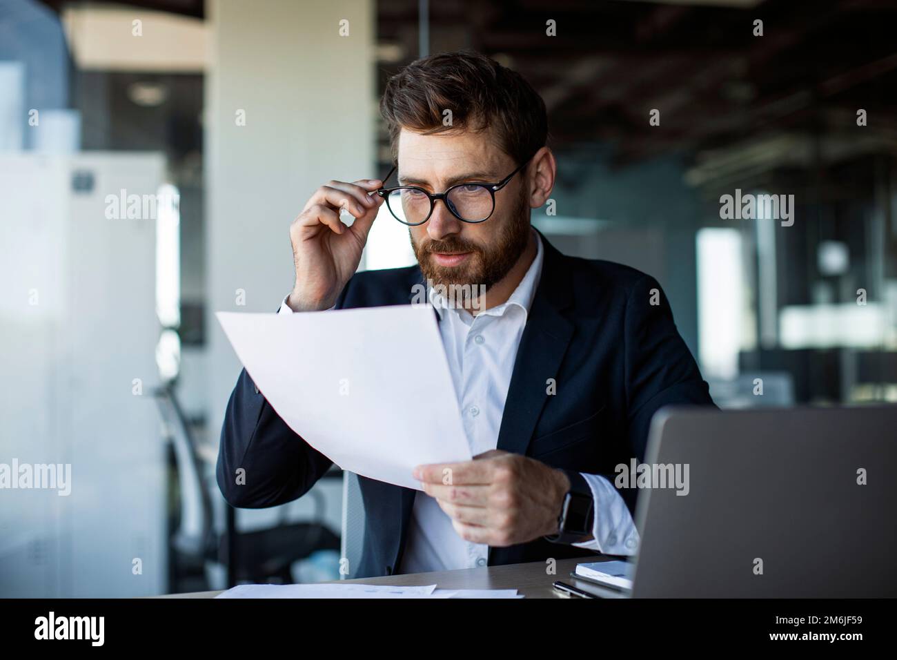 Fokussierter Geschäftsmann mittleren Alters, der mit Papieren in modernen Bürogebäuden arbeitet und Finanzdokumente liest Stockfoto