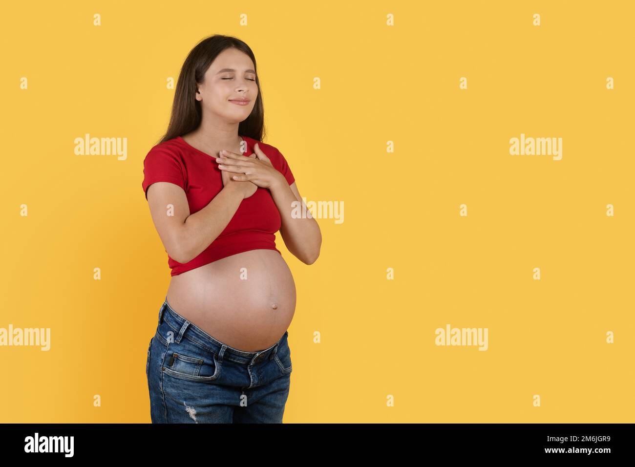 Dankbare Schwangere Frau, Die Die Hände An Der Brust Hält, Während Sie Auf Gelbem Hintergrund Steht Stockfoto