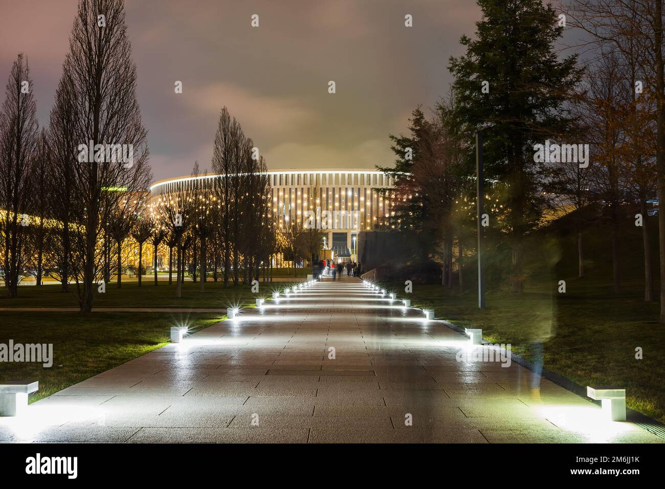 Abendpark mit einem beleuchteten Fußgängerweg, der zu einem halbrunden, großen, leuchtenden Gebäude führt. Stockfoto