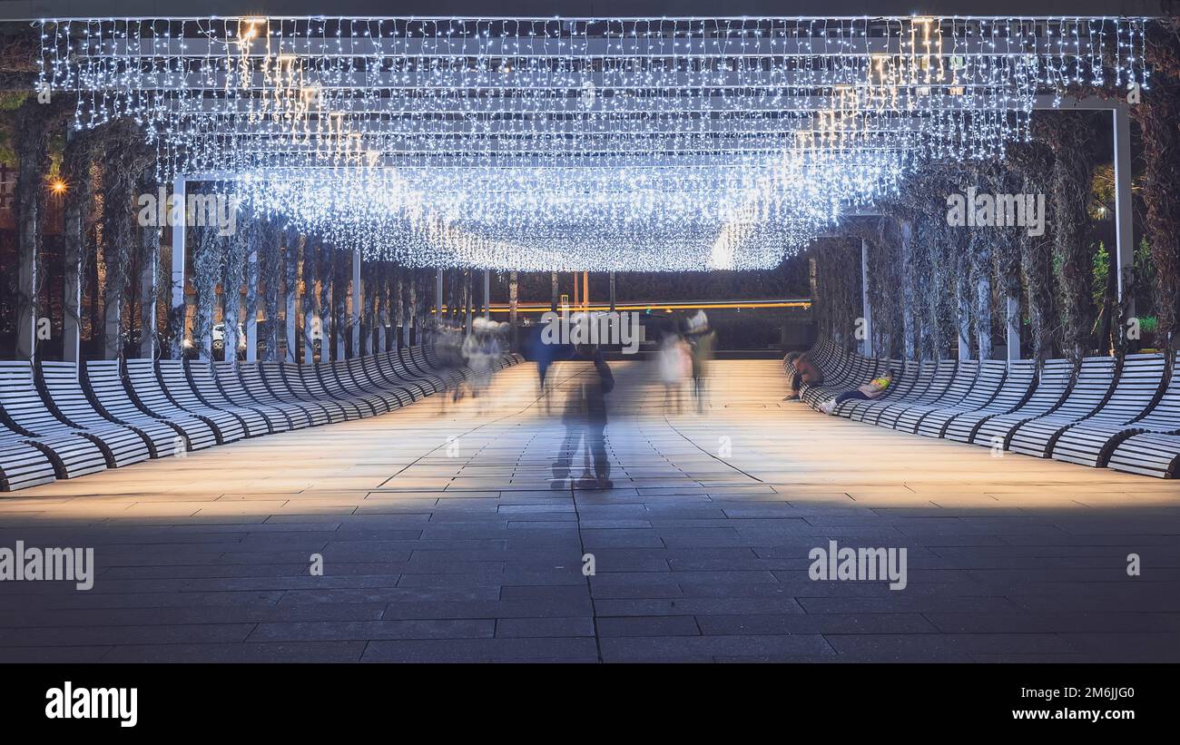 Die Umrisse eines Kindes auf einem Roller unter den vorbeifahrenden Personen entlang der abendlichen Parkgasse, beleuchtet von hängenden Girlanden. Wählen Sie Stockfoto