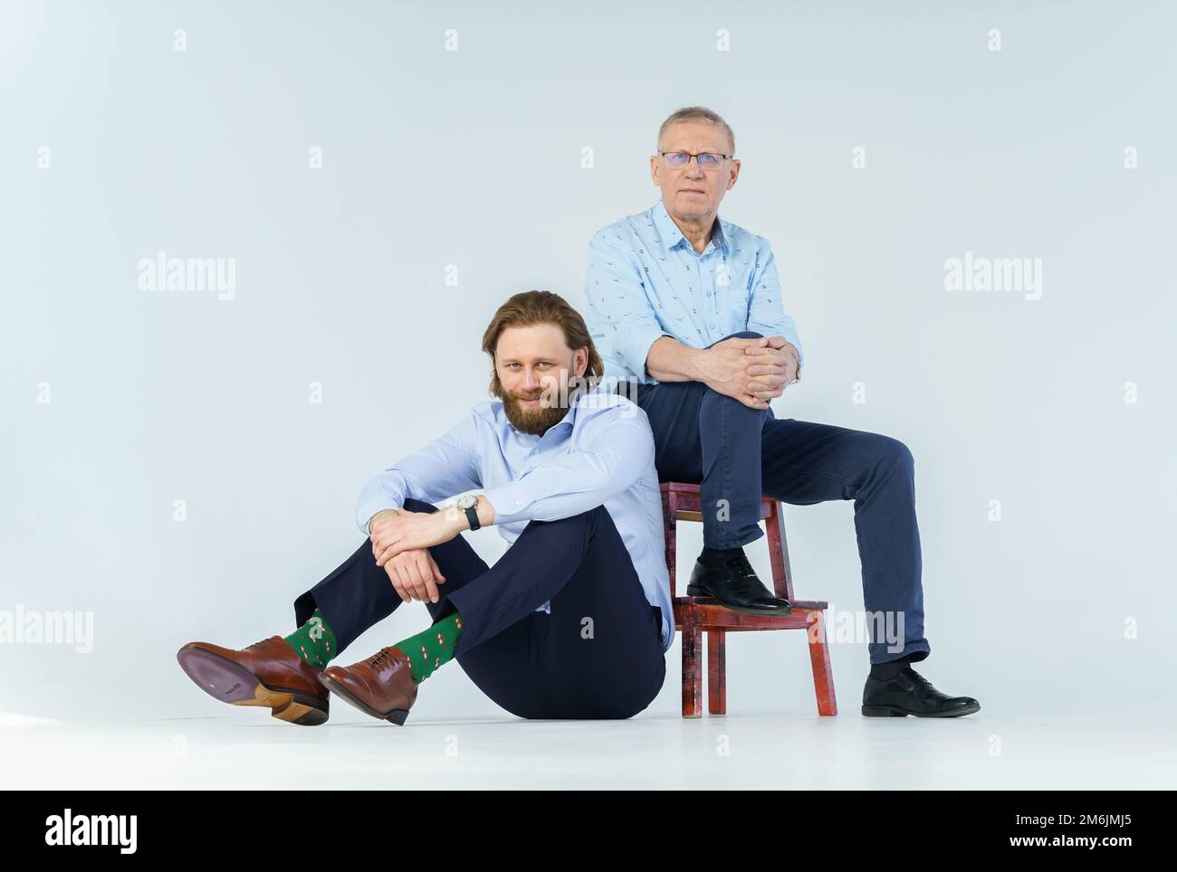 Vater sitzt auf dem Stuhl und Sohn sitzt auf weißem Hintergrund, beide Männer schauen in die Kamera, ein älterer Mann in Brille mit Dioptrien Stockfoto