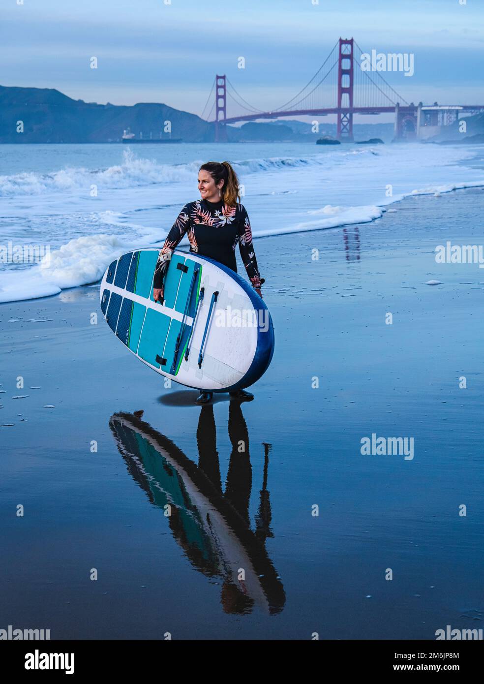 Glückliche Frau mit Paddleboard, die in der Dämmerung am Ufer spaziert Stockfoto