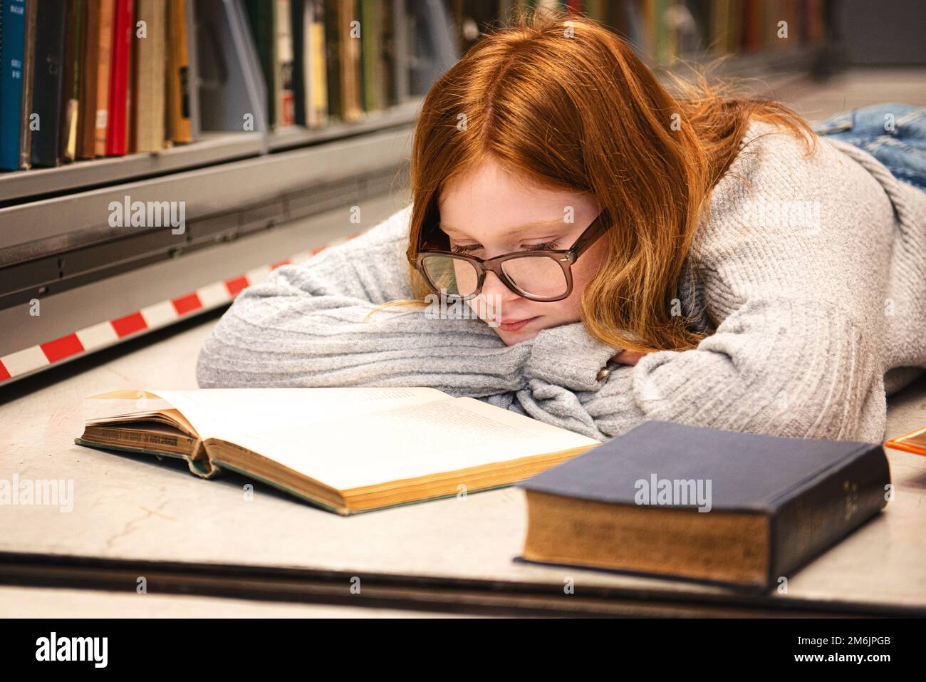 Teenager mit roten Haaren und Brille lag auf dem Boden und las in der Bibliothek. Stockfoto