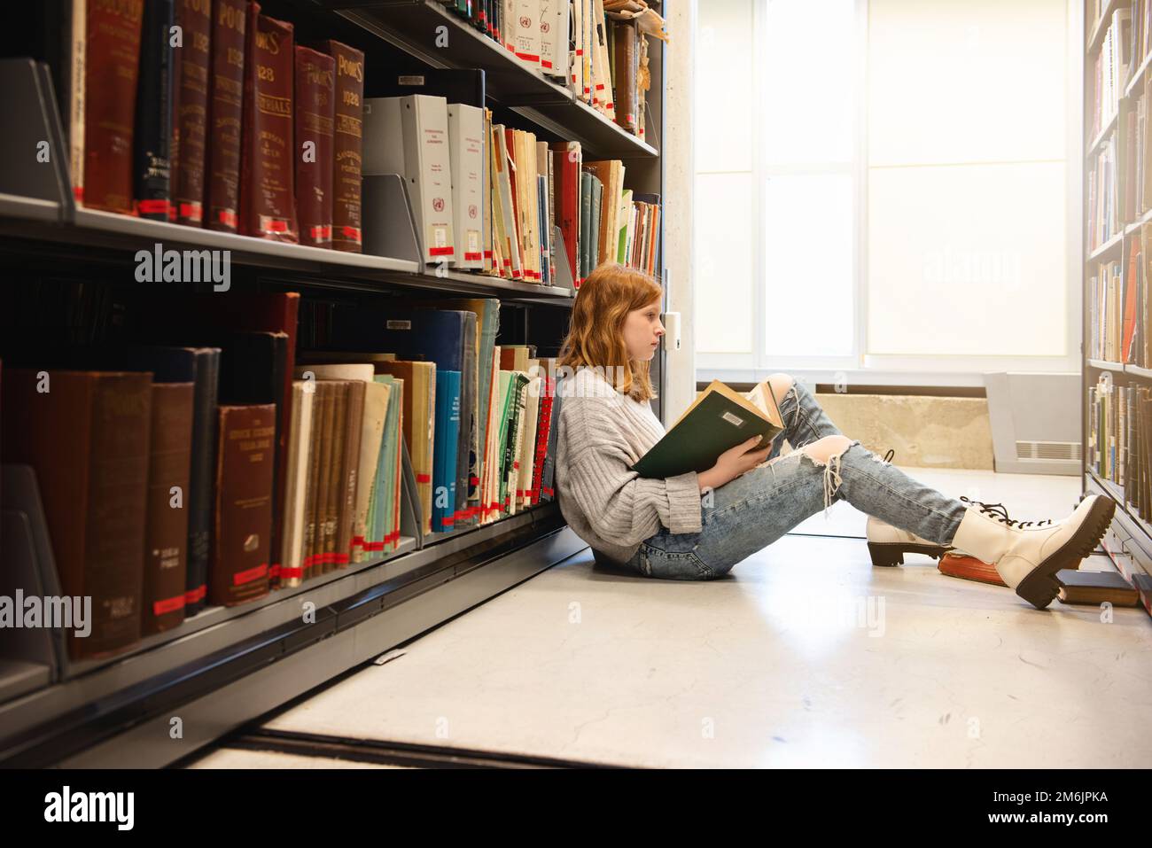 Ein Mädchen mit roten Haaren, das auf dem Boden sitzt und vor Bücherregalen liest. Stockfoto