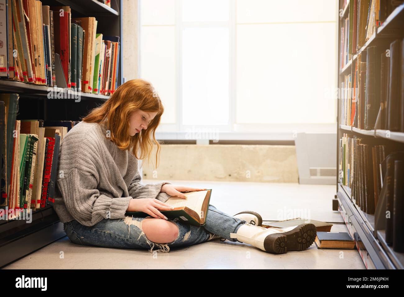 Teenager mit roten Haaren, die ein Buch an Bücherregalen liest. Stockfoto