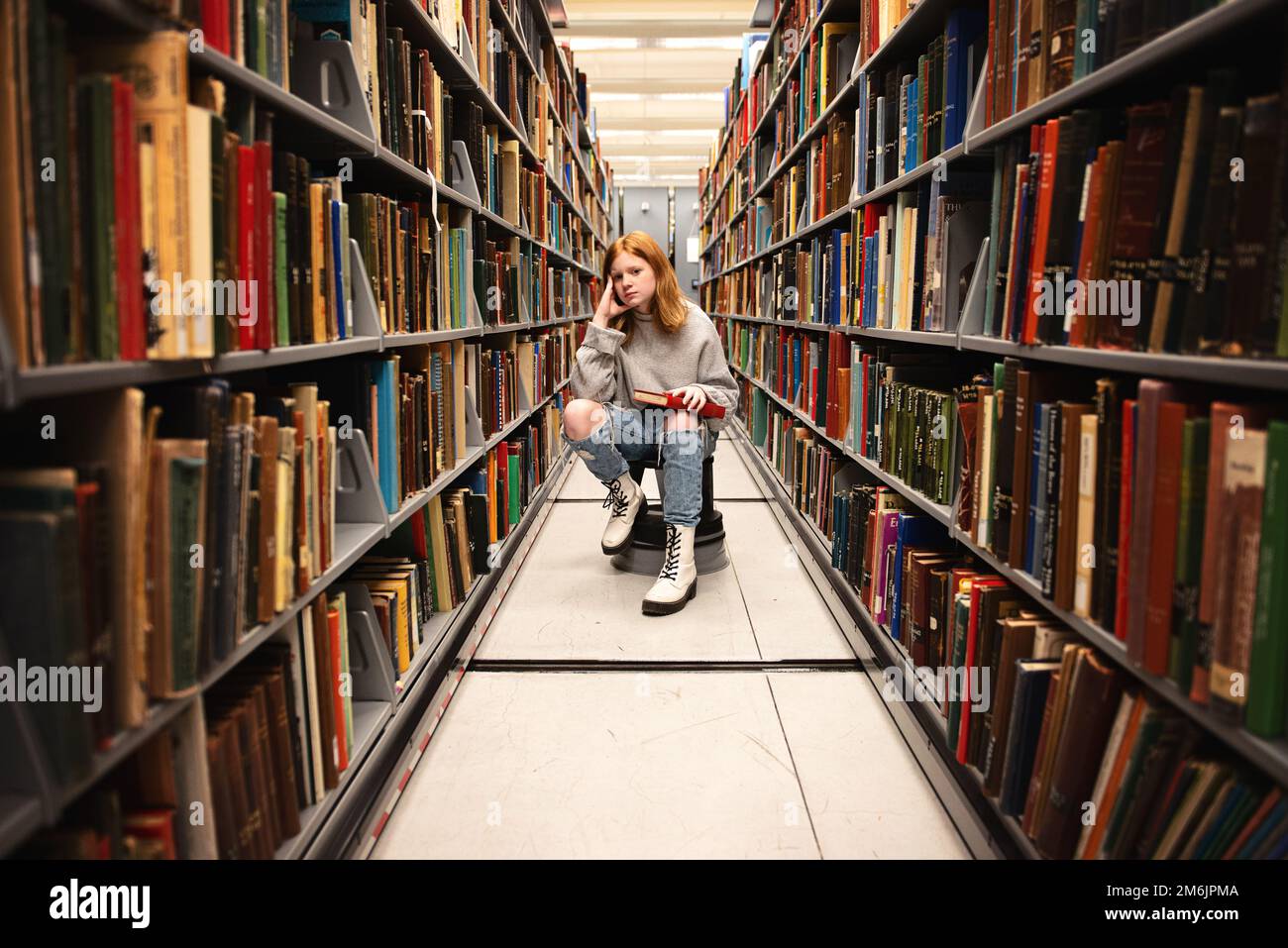 Teenager-Mädchen, die mit Buch zwischen Bücherregalen sitzt. Stockfoto