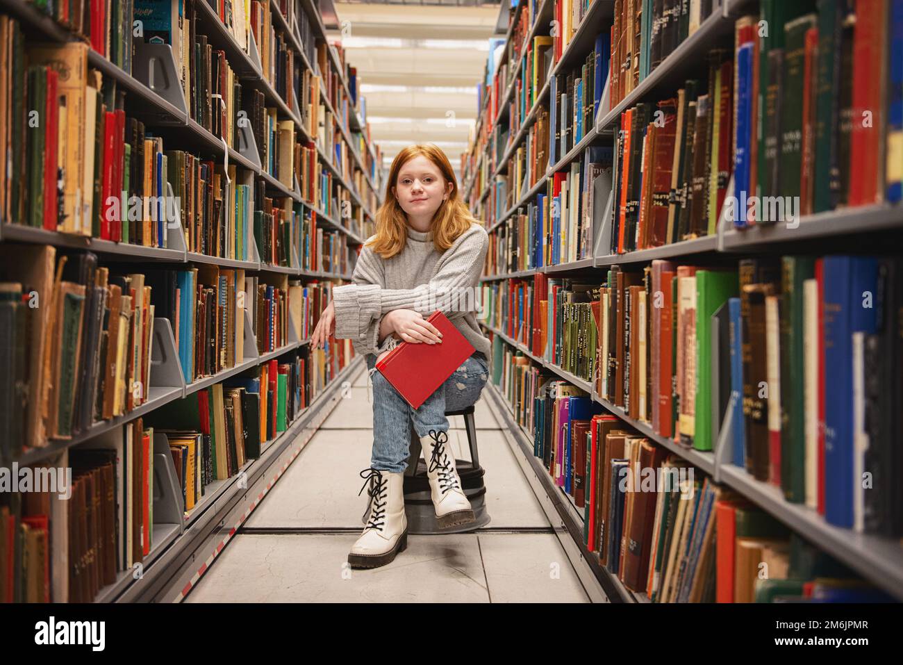 Teenager-Mädchen, die mit Buch zwischen Bücherregalen sitzt. Stockfoto