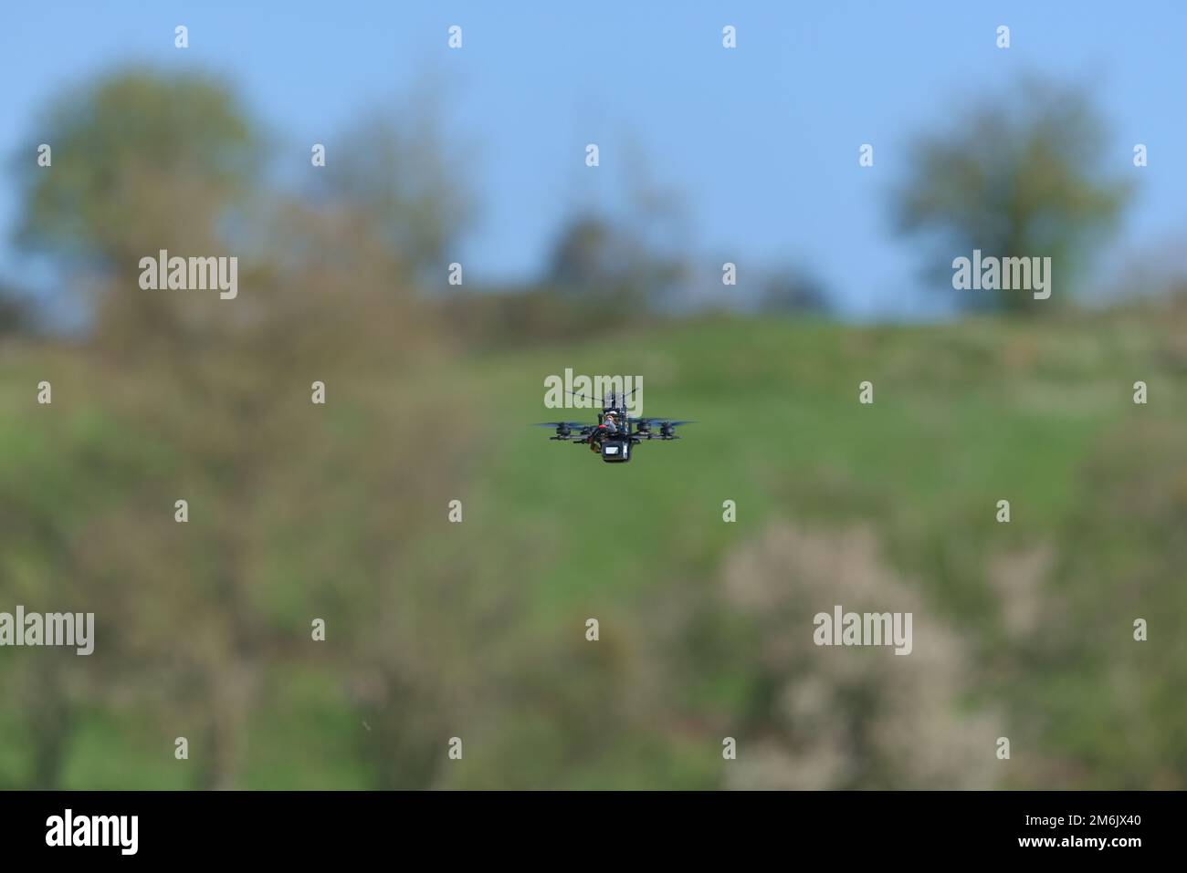 Eine sehr kleine und winzige Zahnstocher-Drohne fliegt von der Kamera weg, in einer ländlichen Umgebung, zu einem von Bäumen gesäumten Horizont und blauem Himmel. Stockfoto