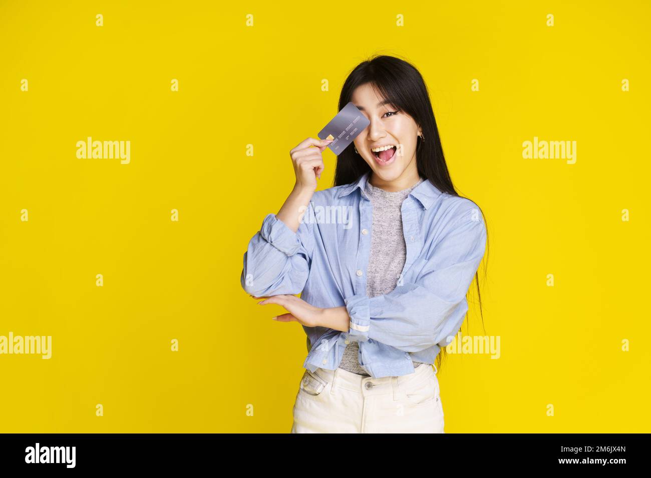 Glückliches asiatisches Mädchen mit Kredit, Debitkarte, die ihr Gesicht berührt, Auge. Asiatisches Mädchen in blauem Hemd, das eine Modell-Kreditkarte hat, isoliert Stockfoto