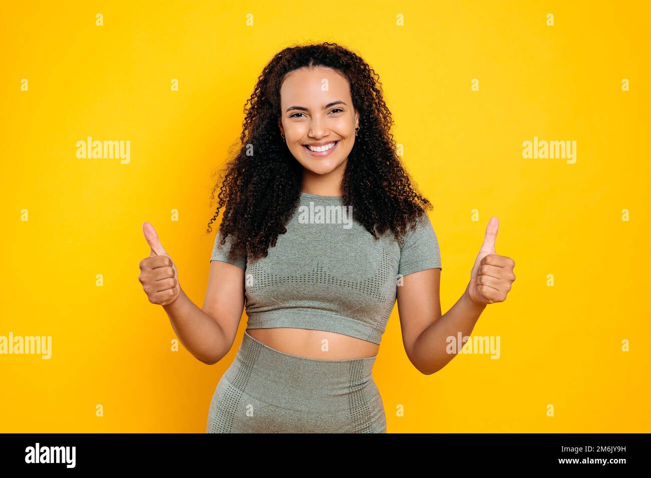 Foto einer positiven, glücklichen, reizenden, lockigen, sportlichen brasilianerin oder hispanischen Sportlerin in Sportbekleidung, mit Daumen-hoch-Geste, sieht in die Kamera, lächelt, steht auf isoliertem orangefarbenen Hintergrund Stockfoto
