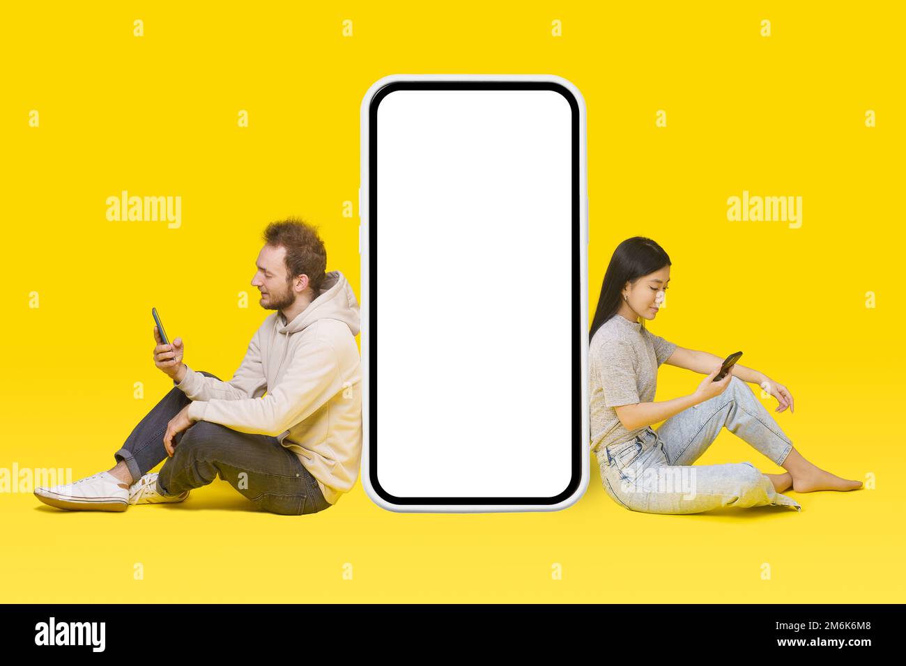 Ein weißer Typ und ein asiatisches Mädchen, die auf dem Boden sitzen, mit Handys in den Händen, lehnen sich auf einen riesigen, riesigen, leeren weißen Smartphone-Bildschirm, Handy A Stockfoto