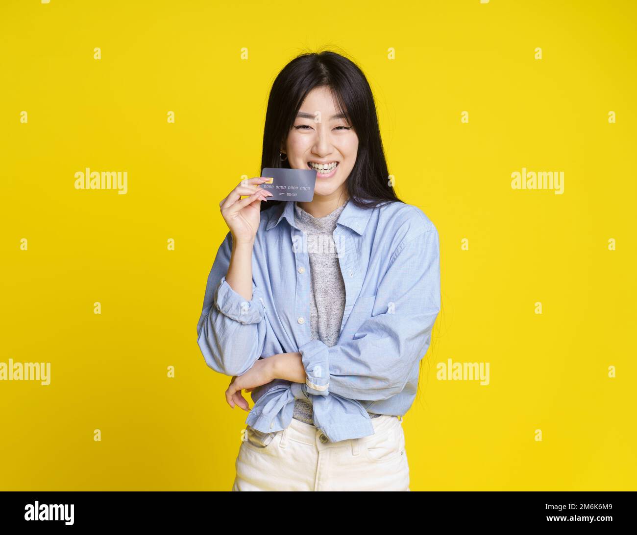 Bankkarte beißen, asiatisches Mädchen mit Kredit- und Debitkarte. Ein asiatisches Mädchen in blauem Hemd, das auf Yello eine Kreditkarte hat Stockfoto