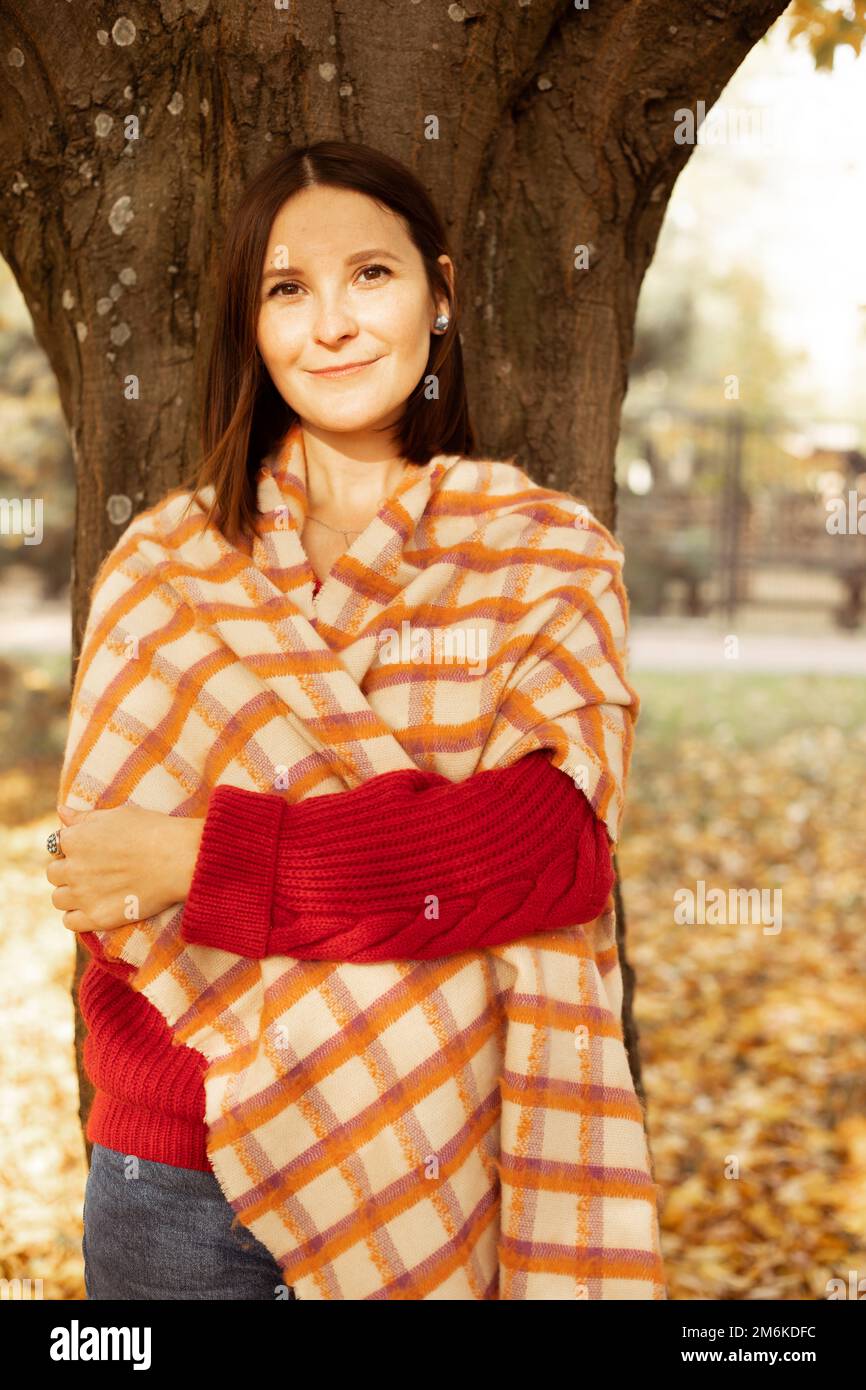 Porträt einer jungen, hübschen Frau, die neben einem Baum steht, mit gekreuzten Händen inmitten gelber gefallener Blätter im Waldpark. Stockfoto