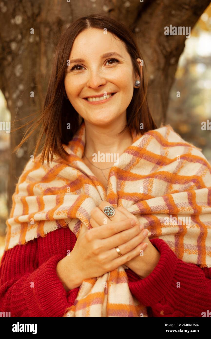 Porträt einer glücklichen jungen, wunderschönen Frau, die einen Schal mit Händen hält, steht neben dem Baum im Waldpark. Vertikal. Herbst. Stockfoto