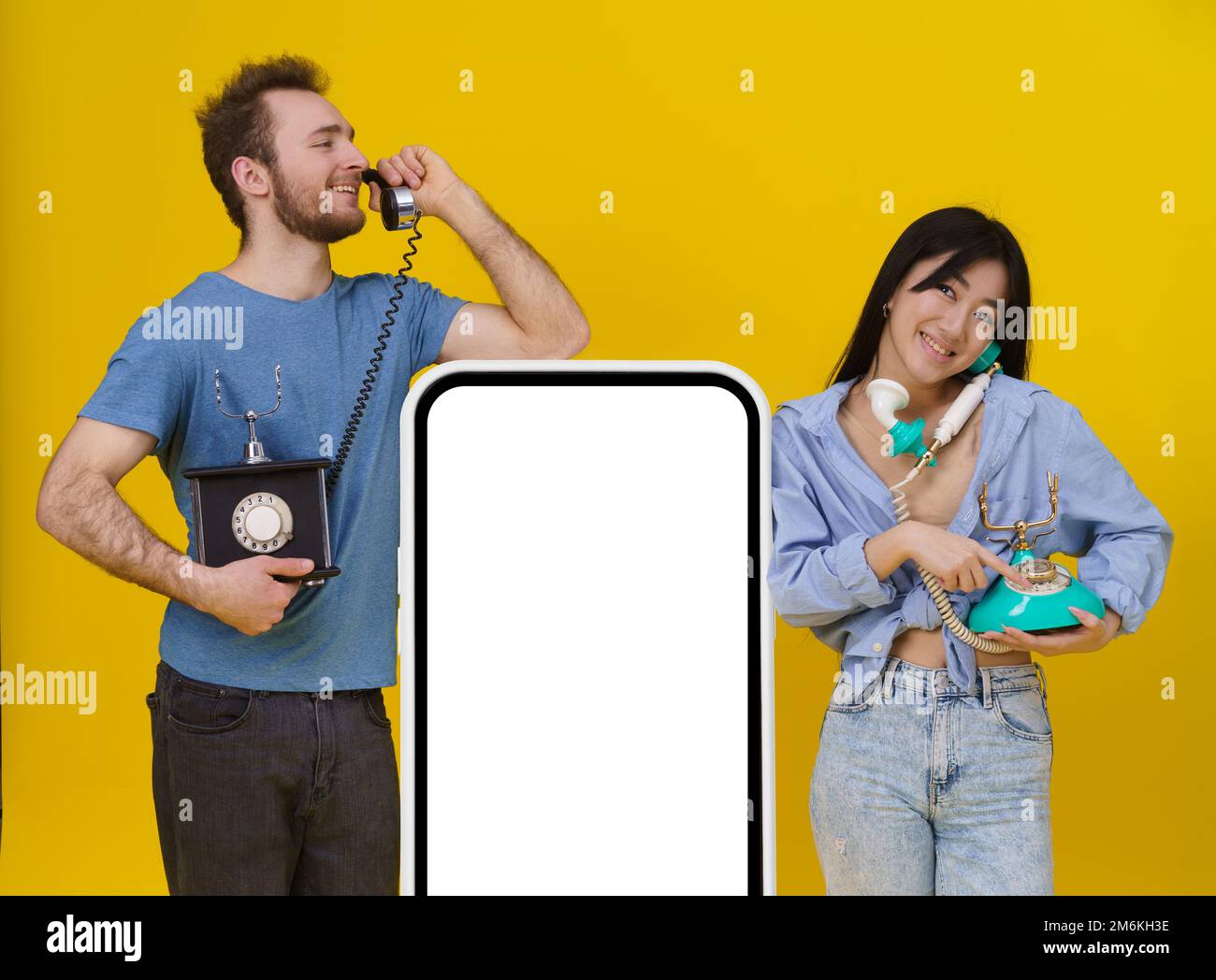 Ein hübscher Kerl und ein asiatisches Mädchen, die sich auf alten Telefonen unterhalten, lehnten sich auf ein riesiges Smartphone oder ein digitales Tablet mit leerem Bildschirm und fröhlichem Smilli Stockfoto