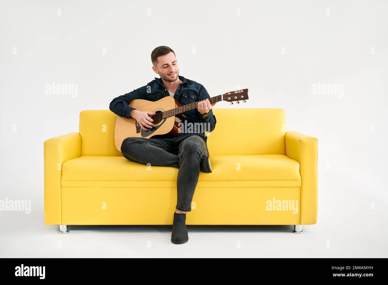 Attraktiver junger Mann, der Akustikgitarre spielt, sitzt auf einem gelben Sofa auf weißem Hintergrund Stockfoto