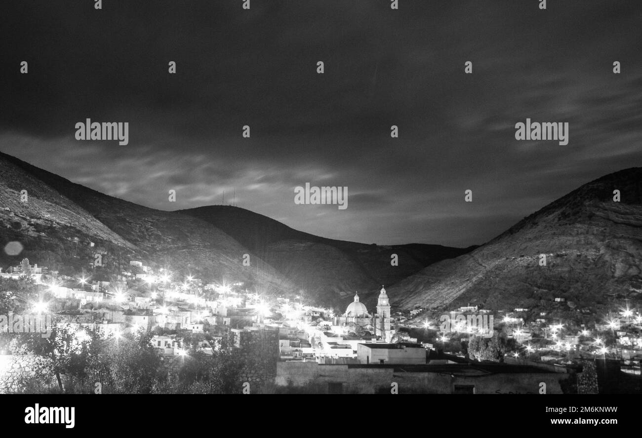 Eine Graustufe der glühenden Real de Catorce Stadtlandschaft bei Nacht, umgeben von hohen Hügeln in Mexiko Stockfoto