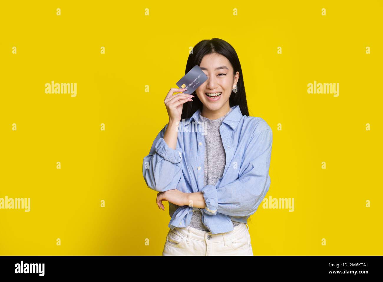 Glückliches, aufgeregtes, junges asiatisches Mädchen mit Kredit- und Debitkarte. Asiatisches Mädchen in blauem Hemd, das eine Modell-Kreditkarte hat, isoliert auf gelbem B. Stockfoto