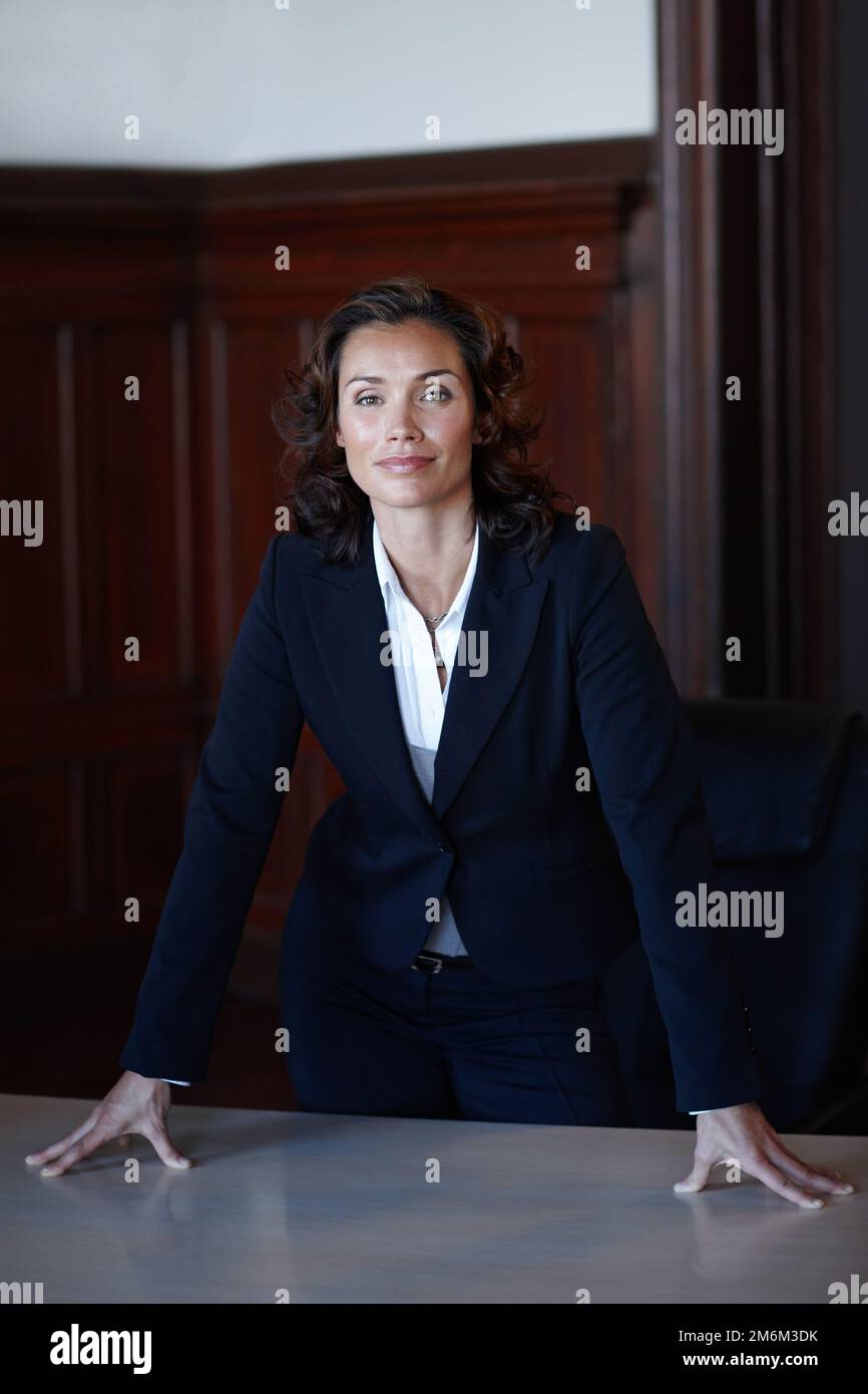 Ich fühle mich mächtig. Attraktive Anwaltsfrau, die sich auf einen Schreibtisch lehnt und in die Kamera lächelt. Stockfoto