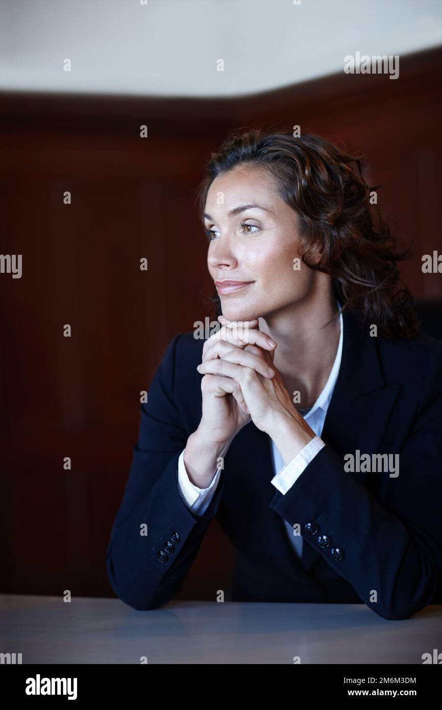 Ich denke über ihren Gerichtsfall nach. Reife Anwaltsfrau, die von der Kamera wegschaut, während sie mit gefalteten Händen sitzt. Stockfoto