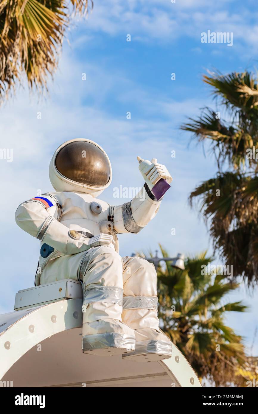 Eine niedliche Figur eines Astronauten in einem Raumanzug, der eine Tube Essen in der Hand hält, sitzt an einem blauen Himmel inmitten von Palmen in einem Stockfoto